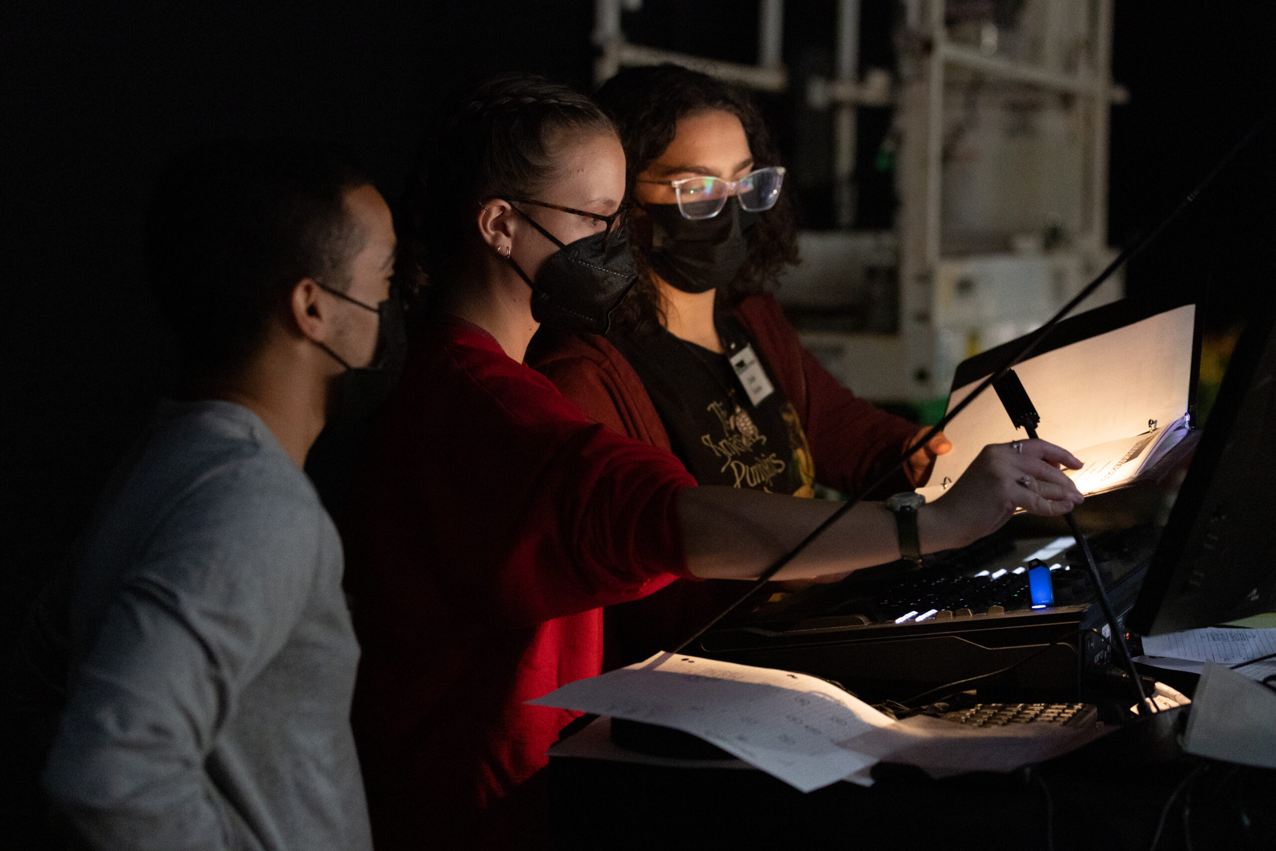 three students in masks looking at a script over a lightboard