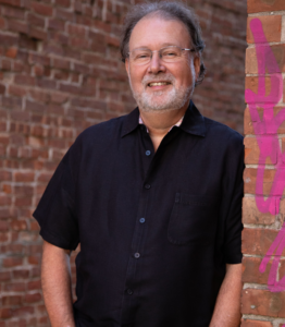 man in black shirt leaning against brick wall and smiling