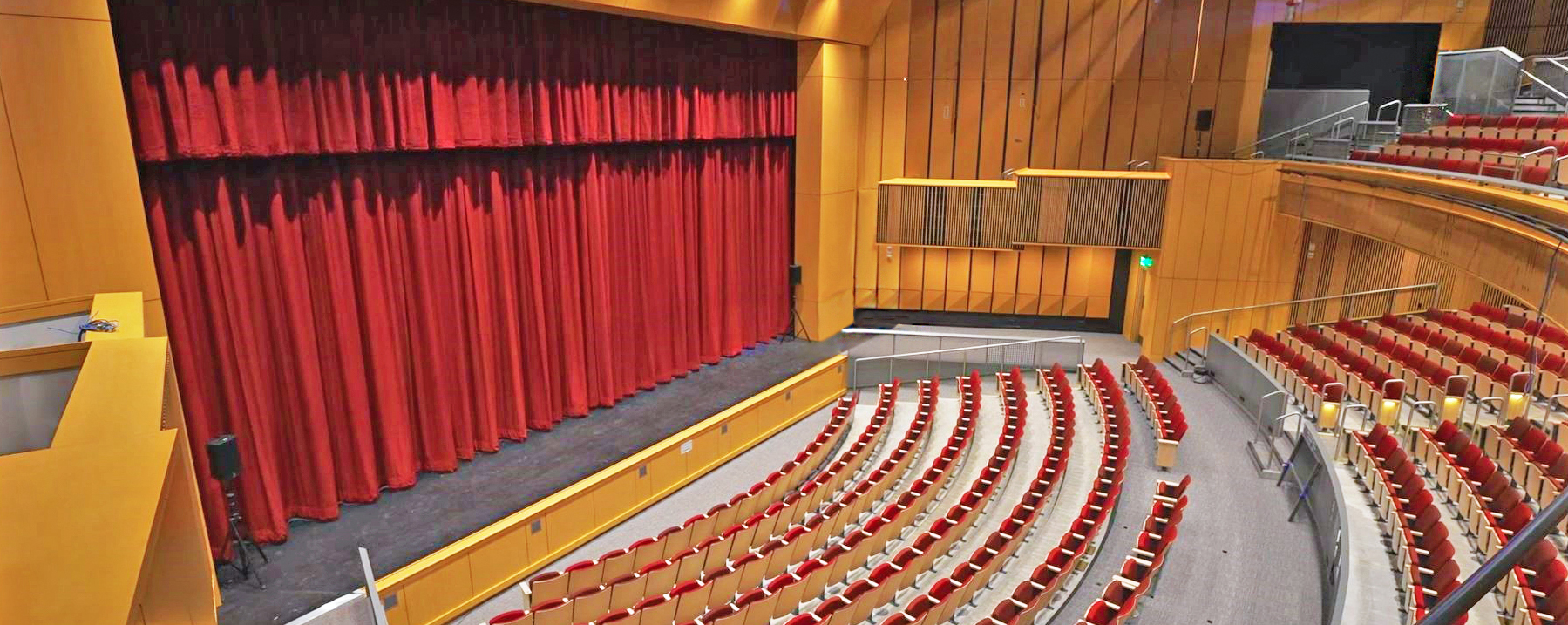 view of the stage and curtain inside an 850-seat auditorium with red seats, a balcony, and wood surroundings