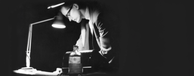 a black and white photo of a man in glasses leaning over a table and drawing lit by a single lamp